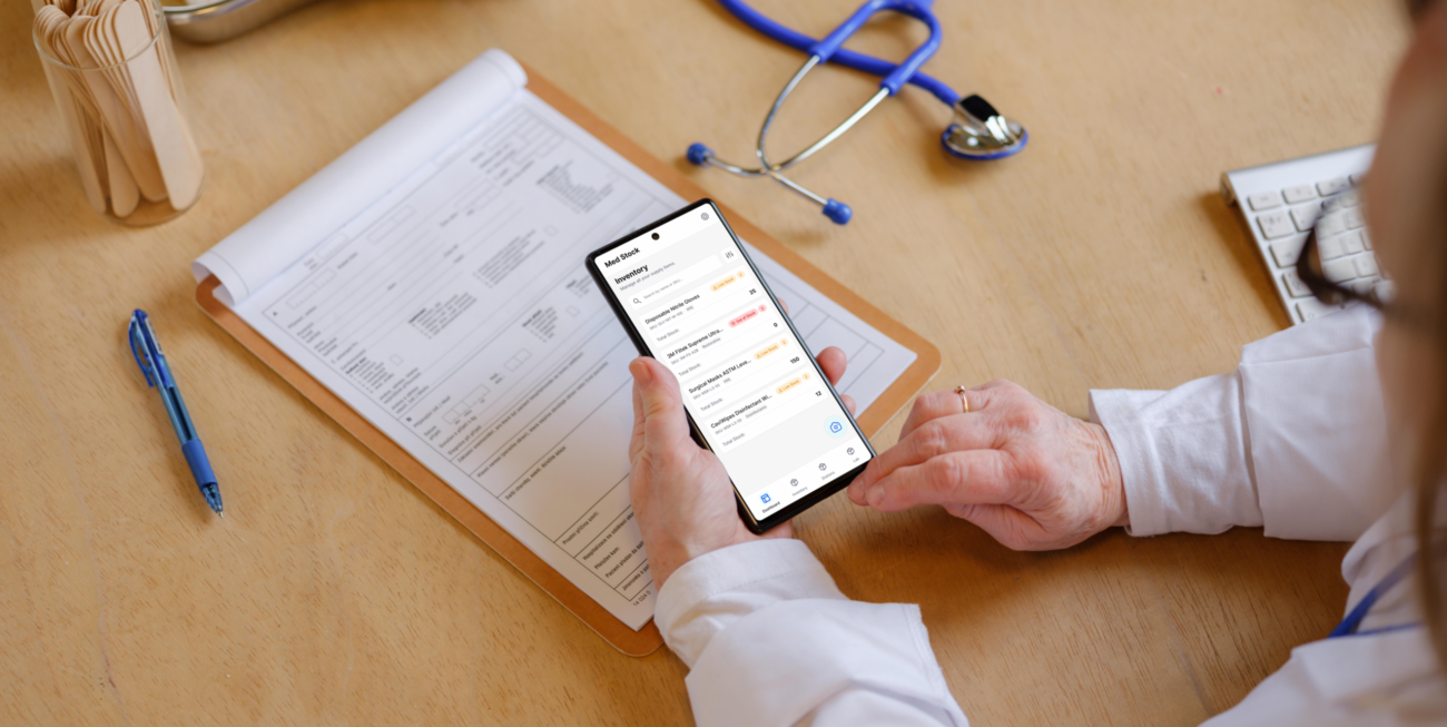 A medical professional in a clinical setting holding a smartphone, actively using the MedStock native app to manage inventory levels alongside traditional medical tools.