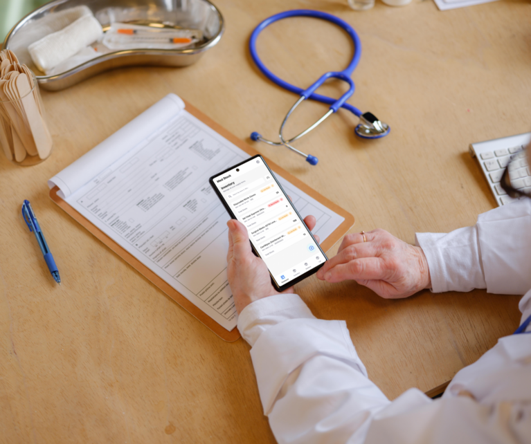 A medical professional in a clinical setting holding a smartphone, actively using the MedStock native app to manage inventory levels alongside traditional medical tools.